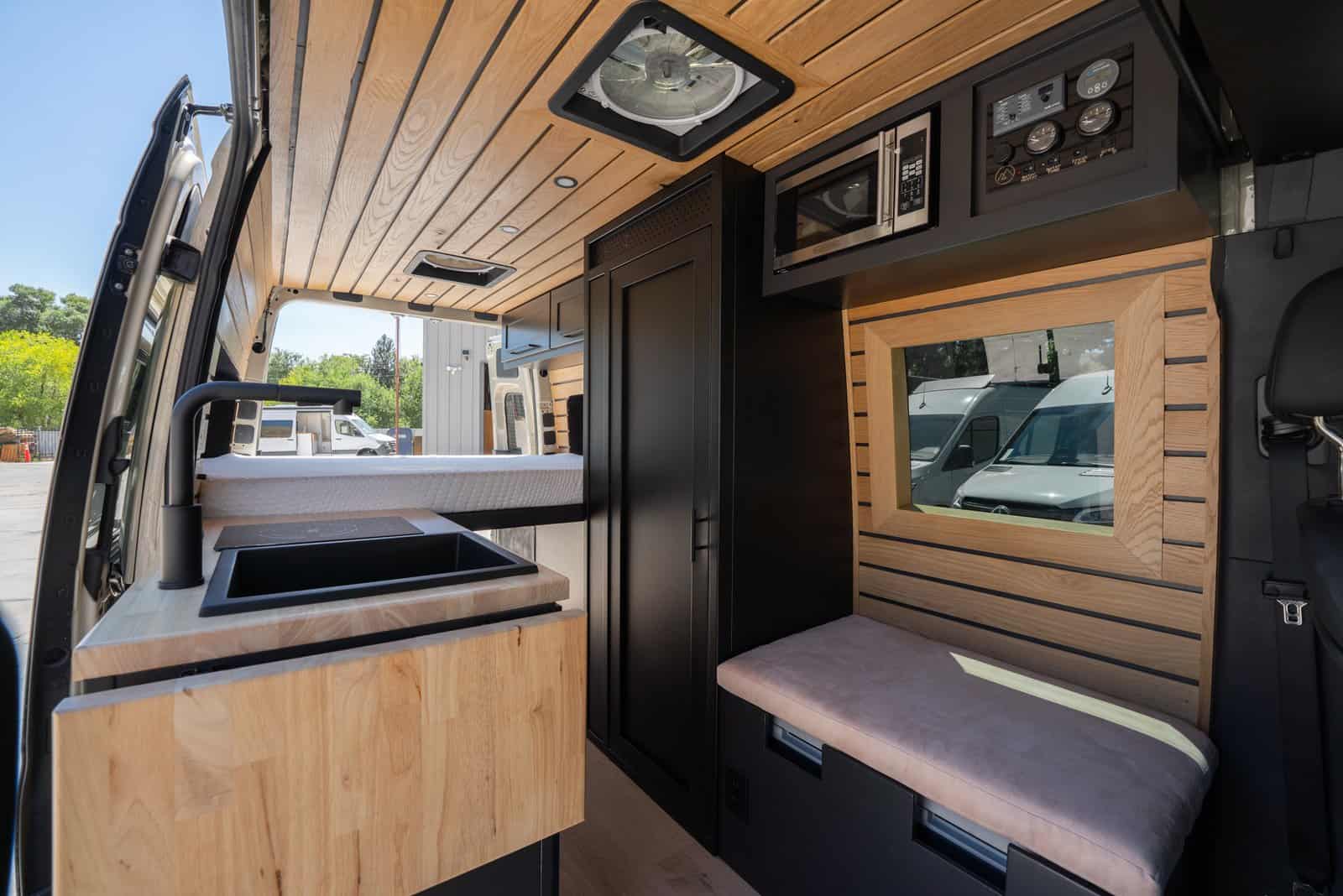 Interior of the camper van with a butcher block galley, black sink, bench seat under a wood-framed window, microwave, and tall black bathroom cabinet