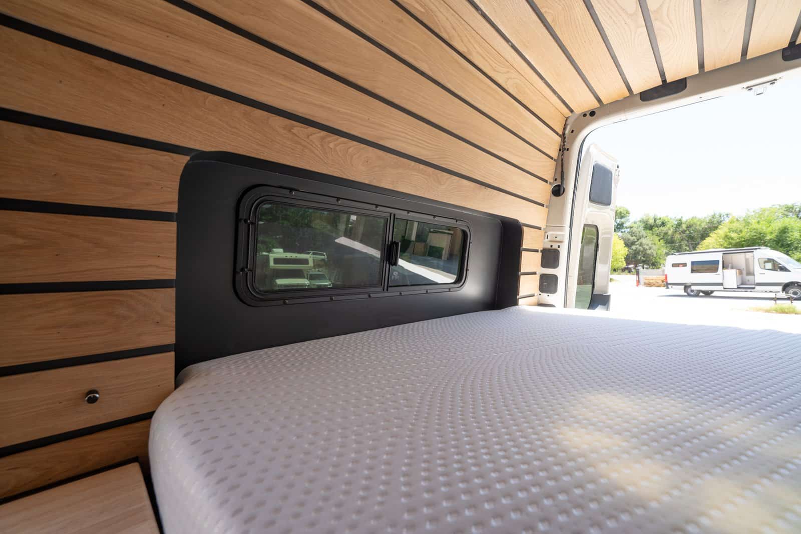 Queen mattress running sideways across the back of the van with a window, wood slat walls, and overhead cabinets