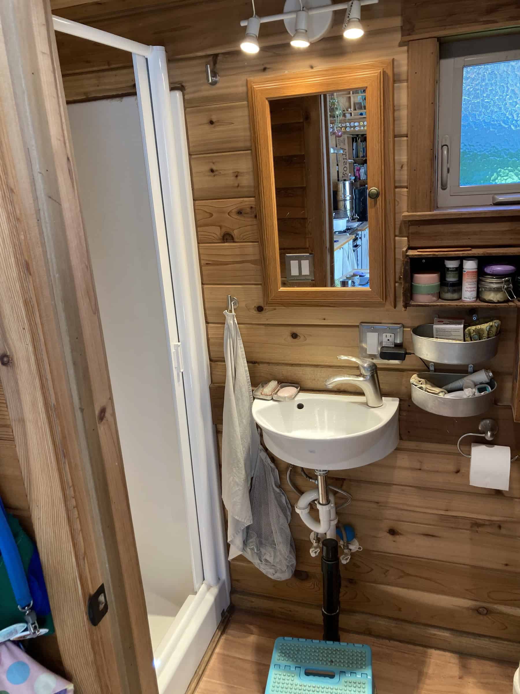 Compact wood-paneled bathroom with a wall-mounted white sink, a wood-framed medicine cabinet, metal basket shelves, and the edge of a white shower stall
