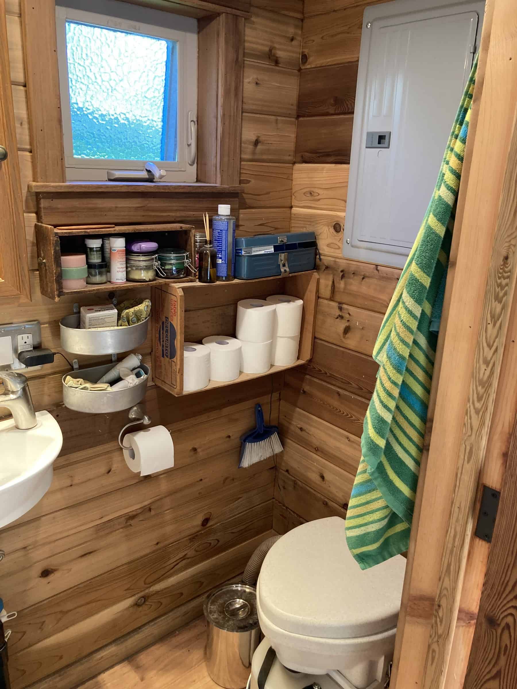 Nature's Head composting toilet in a wood-paneled bathroom with crate shelves of supplies, a small window, and a grey vent hose