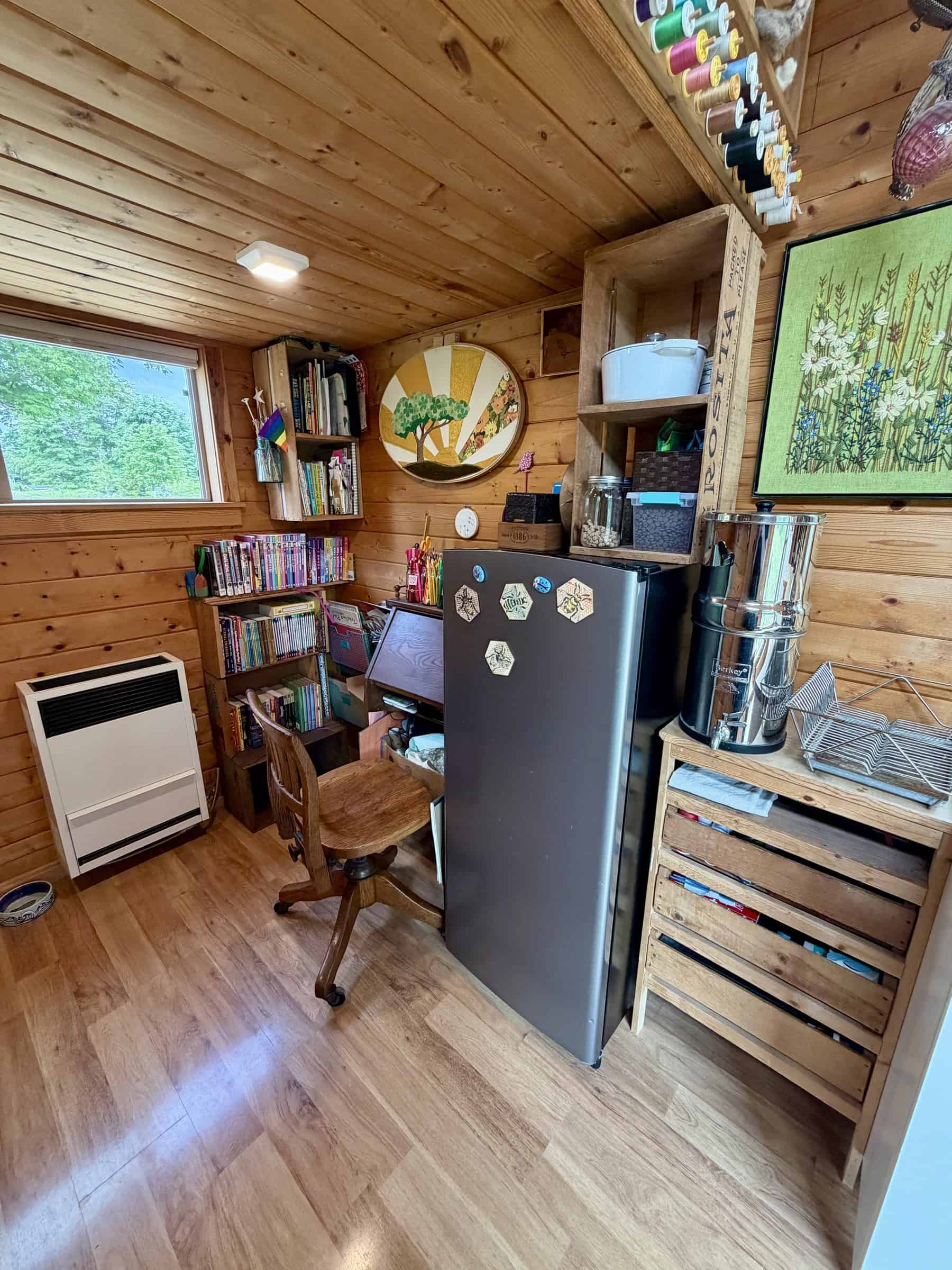 Corner with a propane wall heater under a window, a small wood desk and chair, overflowing crate bookshelves, and a slate apartment-size refrigerator