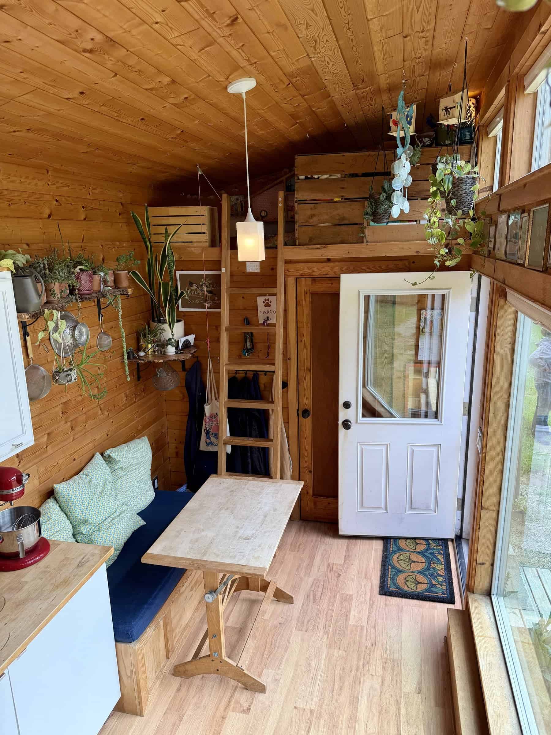 Built-in wood storage bench with a navy cushion and a small table, plants on a live-edge shelf, a ladder to a twin loft, and a white glass entry door