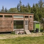 Wood-sided 8 by 32 foot tiny house on a trailer in a grassy Port Angeles clearing, with a large picture window, a glass entry door, and a ground-mounted solar array beside a small green shed