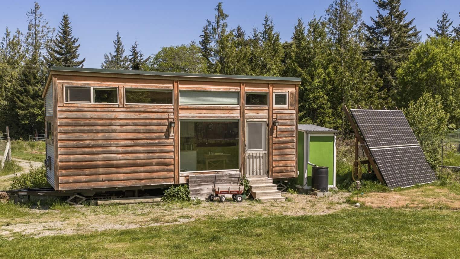 Wood-sided 8 by 32 foot tiny house on a trailer in a grassy Port Angeles clearing, with a large picture window, a glass entry door, and a ground-mounted solar array beside a small green shed