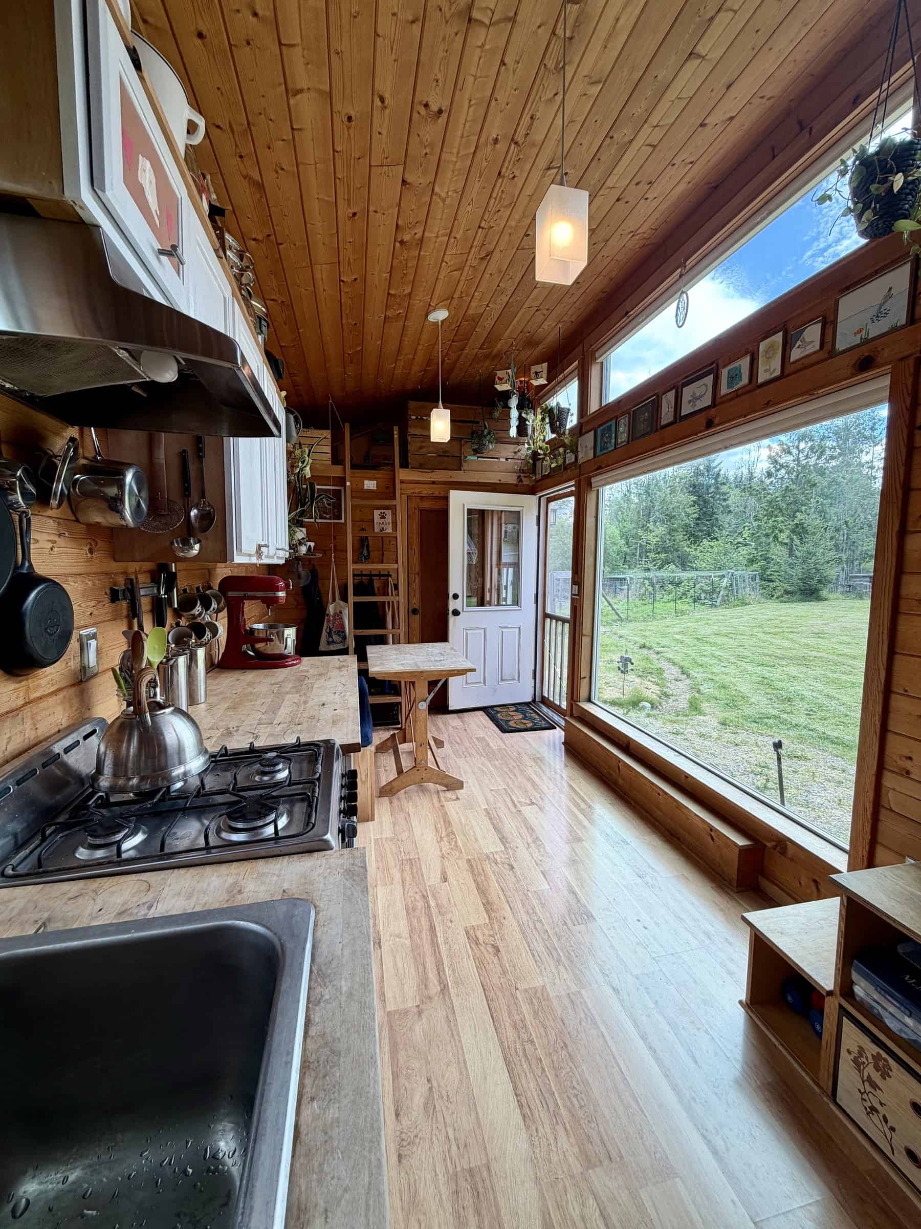 View from a loft toward the kitchen sink and propane range, a small wood table, and the large picture window looking onto a green field