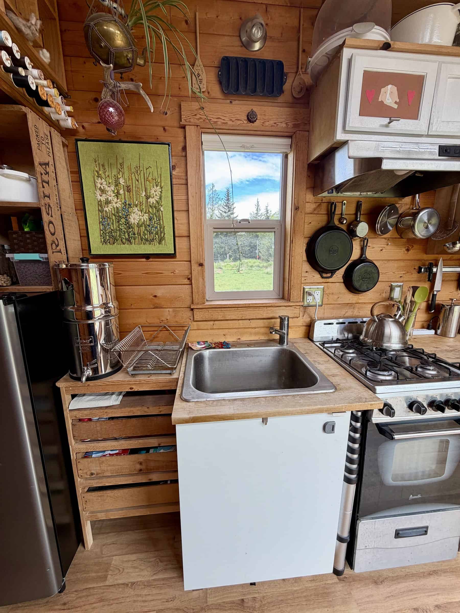 Compact wood-paneled kitchen with a stainless sink under a window, a four-burner propane range and oven, hanging cast-iron pans, and a gravity water filter