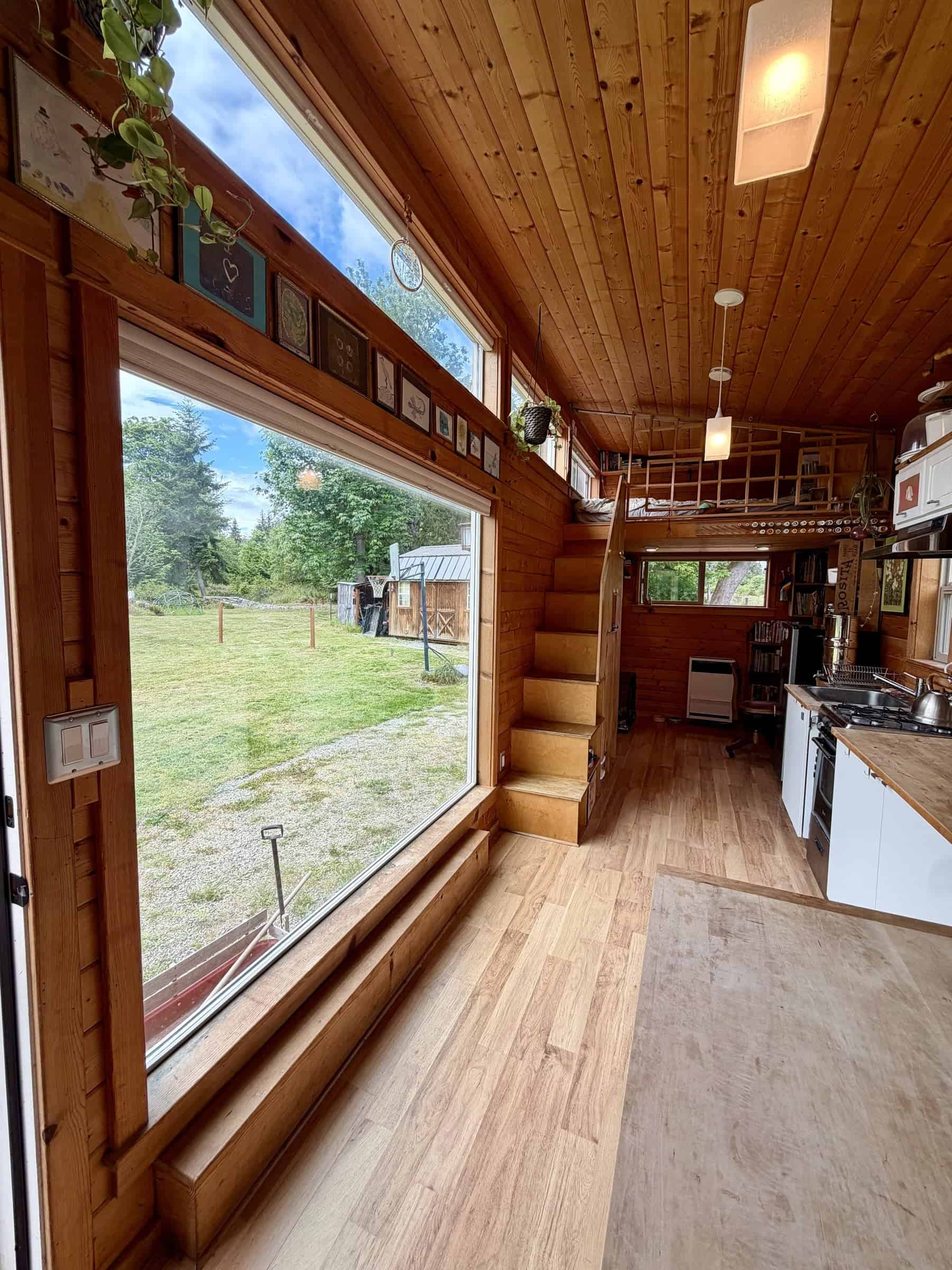 Looking down the wood-paneled tiny house: a huge picture window on the left, storage stairs to a loft, white kitchen cabinets and a propane wall heater under the loft