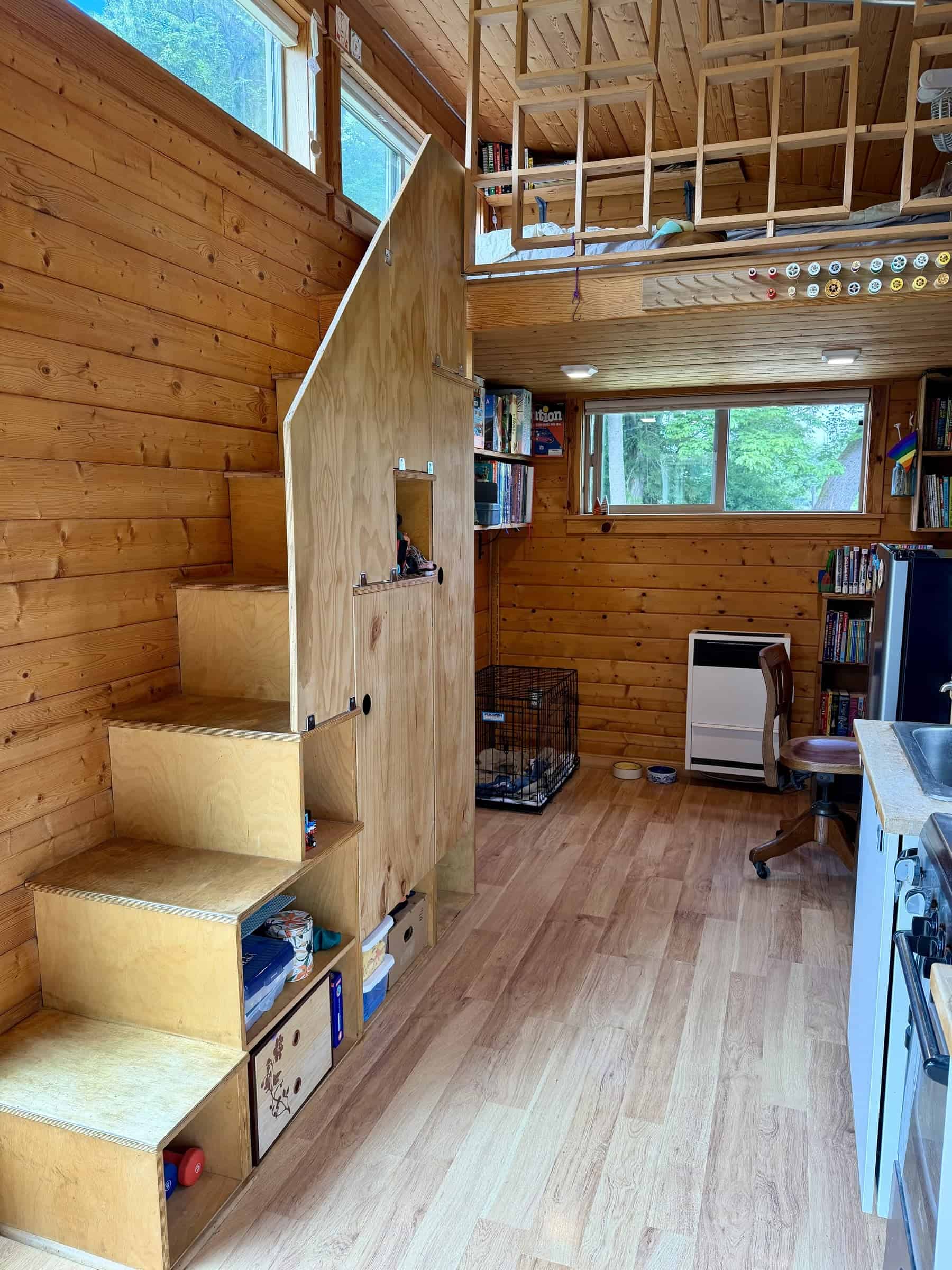 Plywood storage stairs with cubbies and cabinet doors climbing to a loft with a square-grid wood railing, a propane heater and bookshelves below