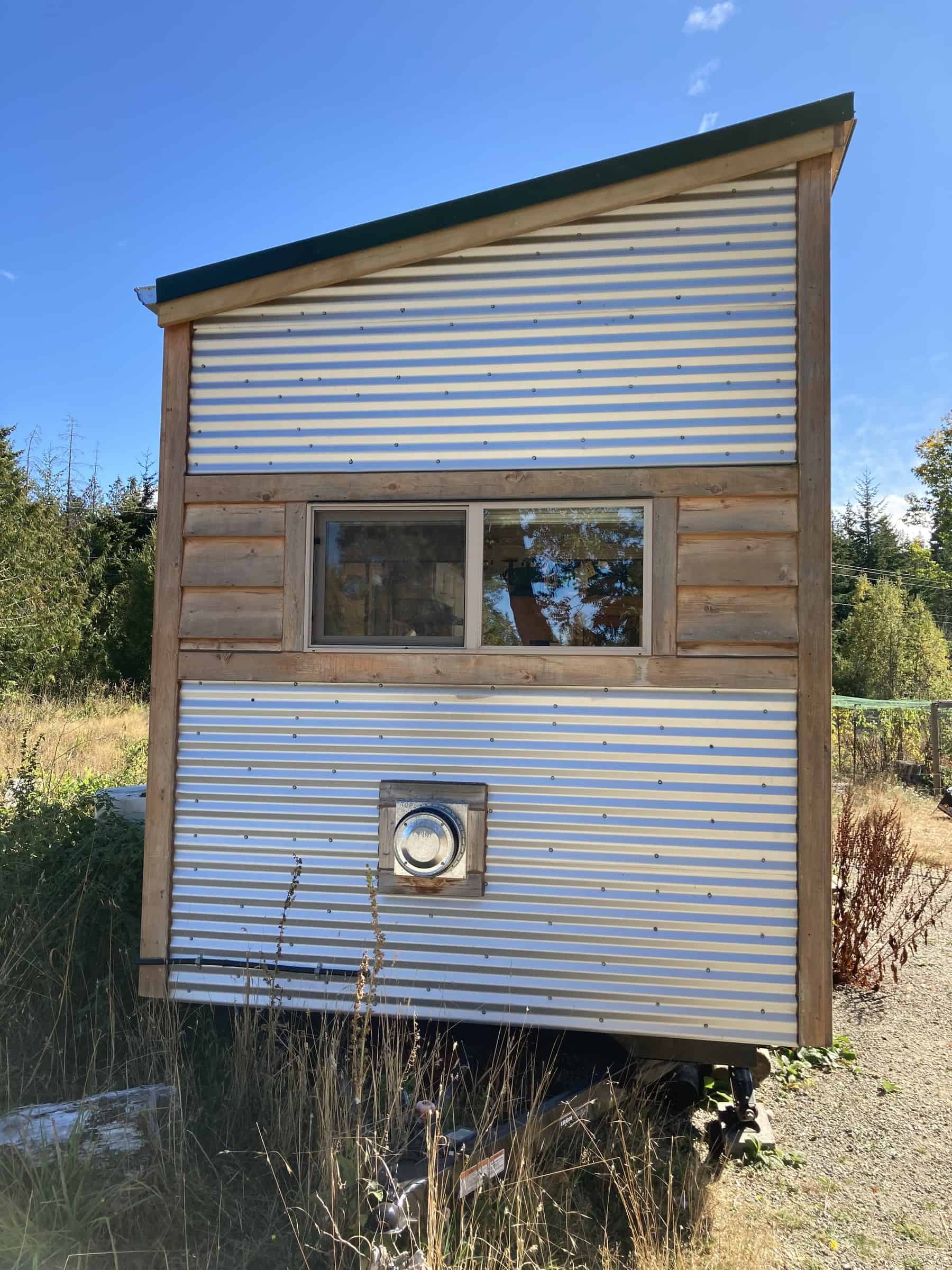 Hitch end of the tiny house on a black trailer, with corrugated metal and a band of wood siding around a sliding window and a circular vent