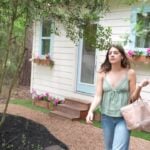 Annie walking past her cream tiny house with light blue window trim and pink flower boxes in the woods