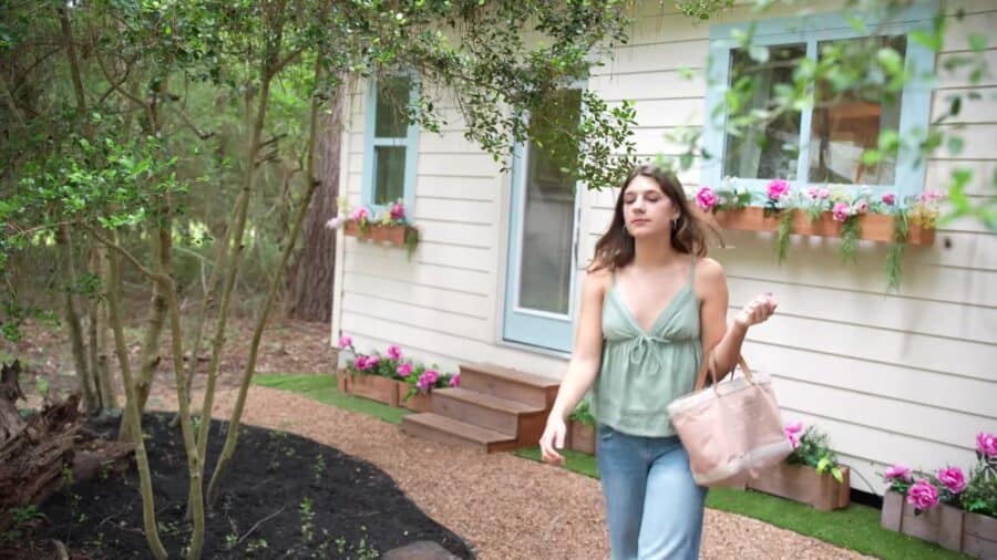 Annie walking past her cream tiny house with light blue window trim and pink flower boxes in the woods