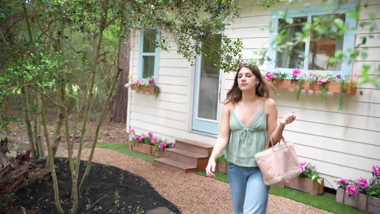 Annie walking past her cream tiny house with light blue window trim and pink flower boxes in the woods