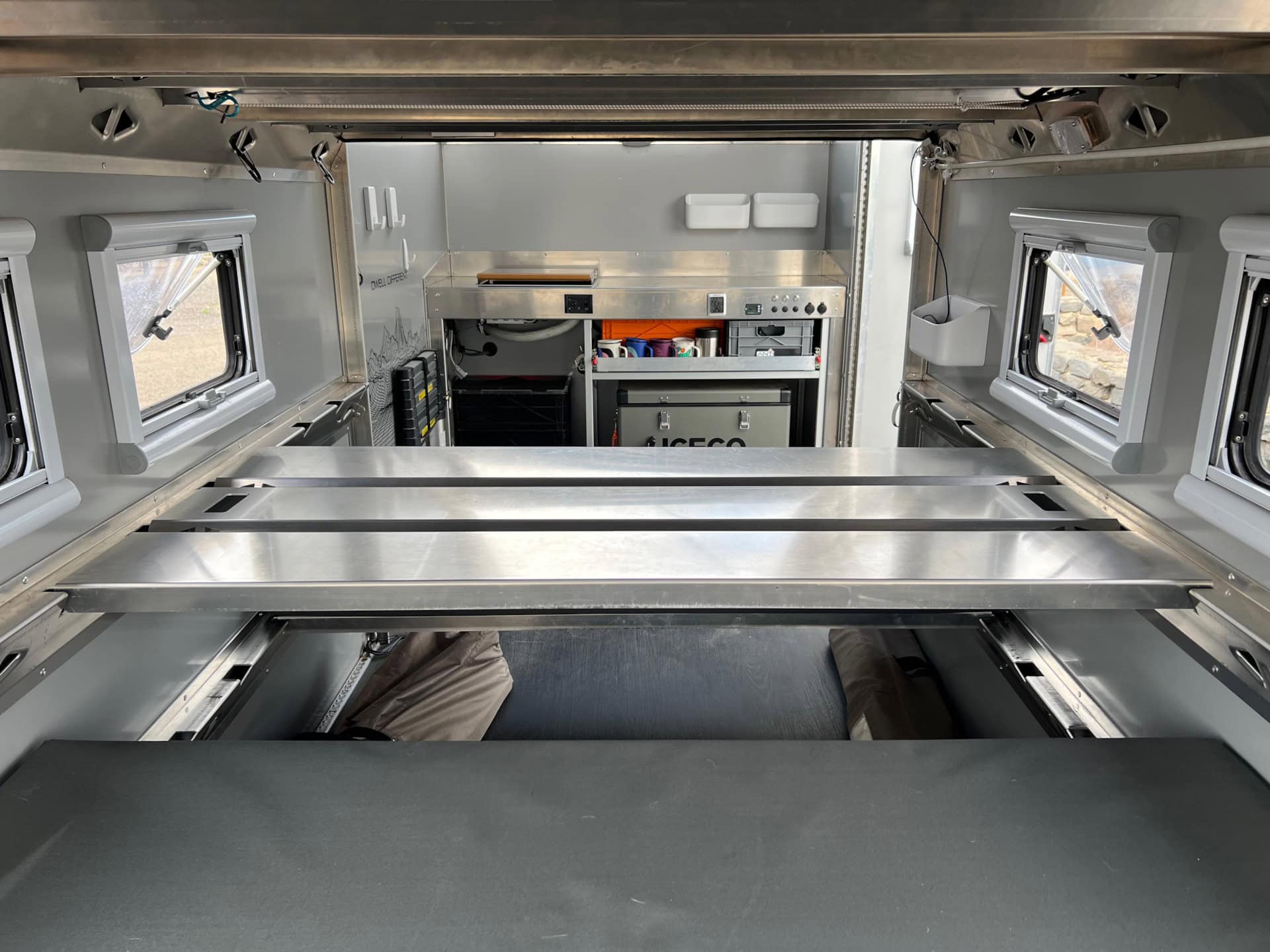 Interior looking forward through aluminum bunk slats toward the kitchen module, ICECO fridge, and windows