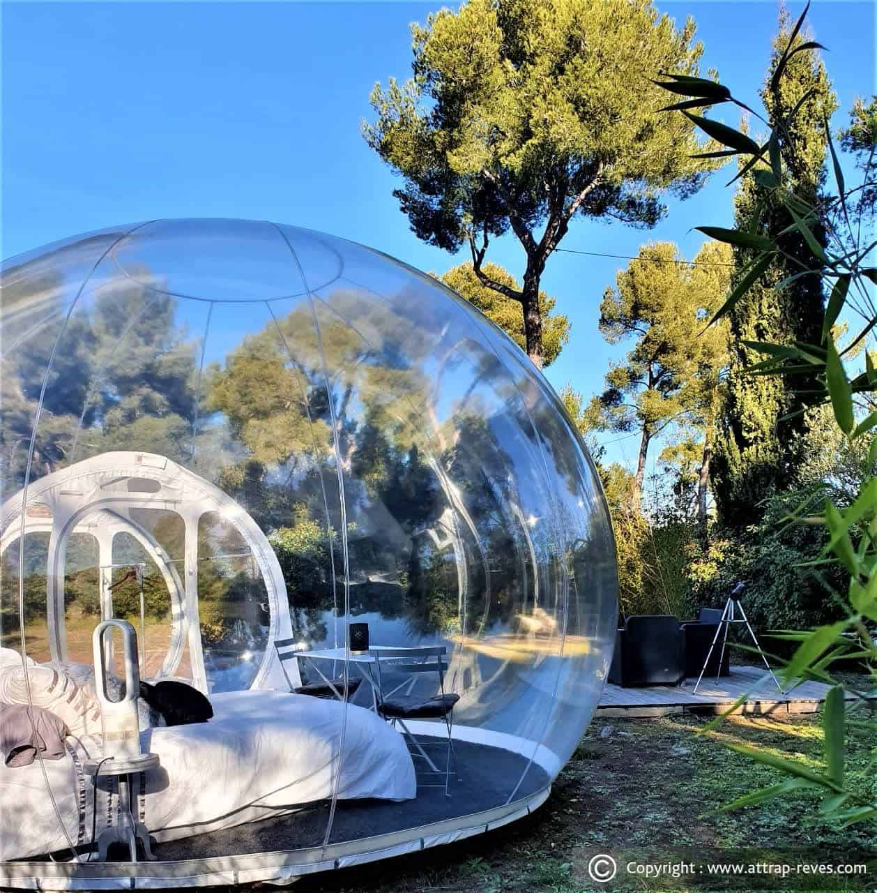 Clear bubble bedroom with white linens beside a wooden deck holding outdoor chairs and a telescope