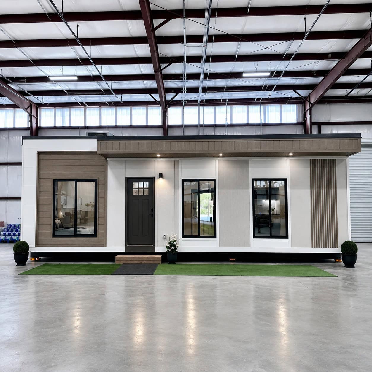 Front of a flat-roof white and tan cottage inside a factory, black windows and a dark door