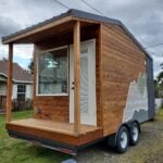 Backcountry The Traveler on a dual-axle trailer: cedar siding, dark metal at the rear, covered porch with wood posts, mountain graphic on the side, Hampstead New Hampshire