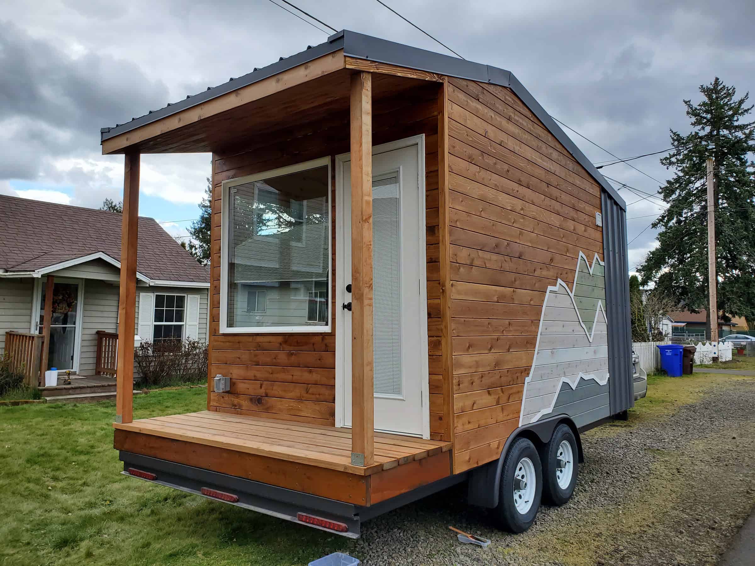 Backcountry The Traveler on a dual-axle trailer: cedar siding, dark metal at the rear, covered porch with wood posts, mountain graphic on the side, Hampstead New Hampshire