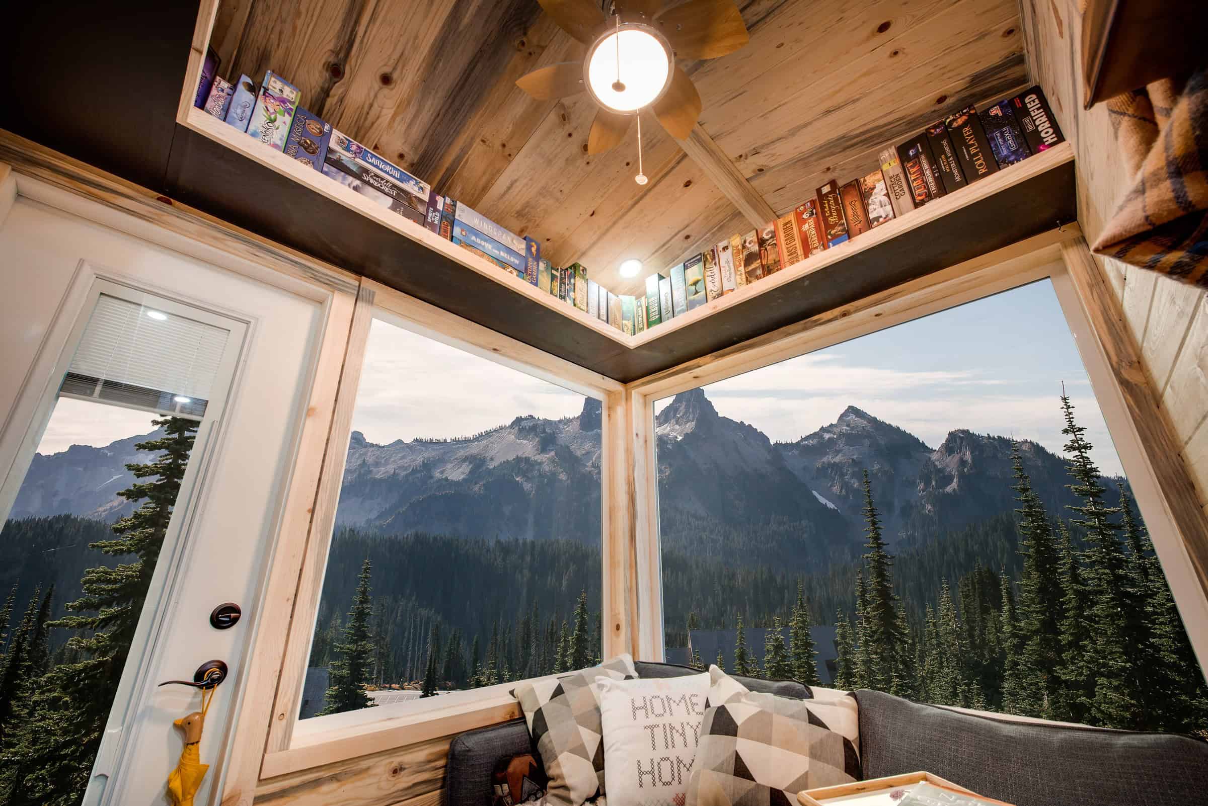 Traveler living corner: two large windows meeting at 90 degrees, wrap-around shelf of games, grey sofa, knotty pine walls and vaulted ceiling