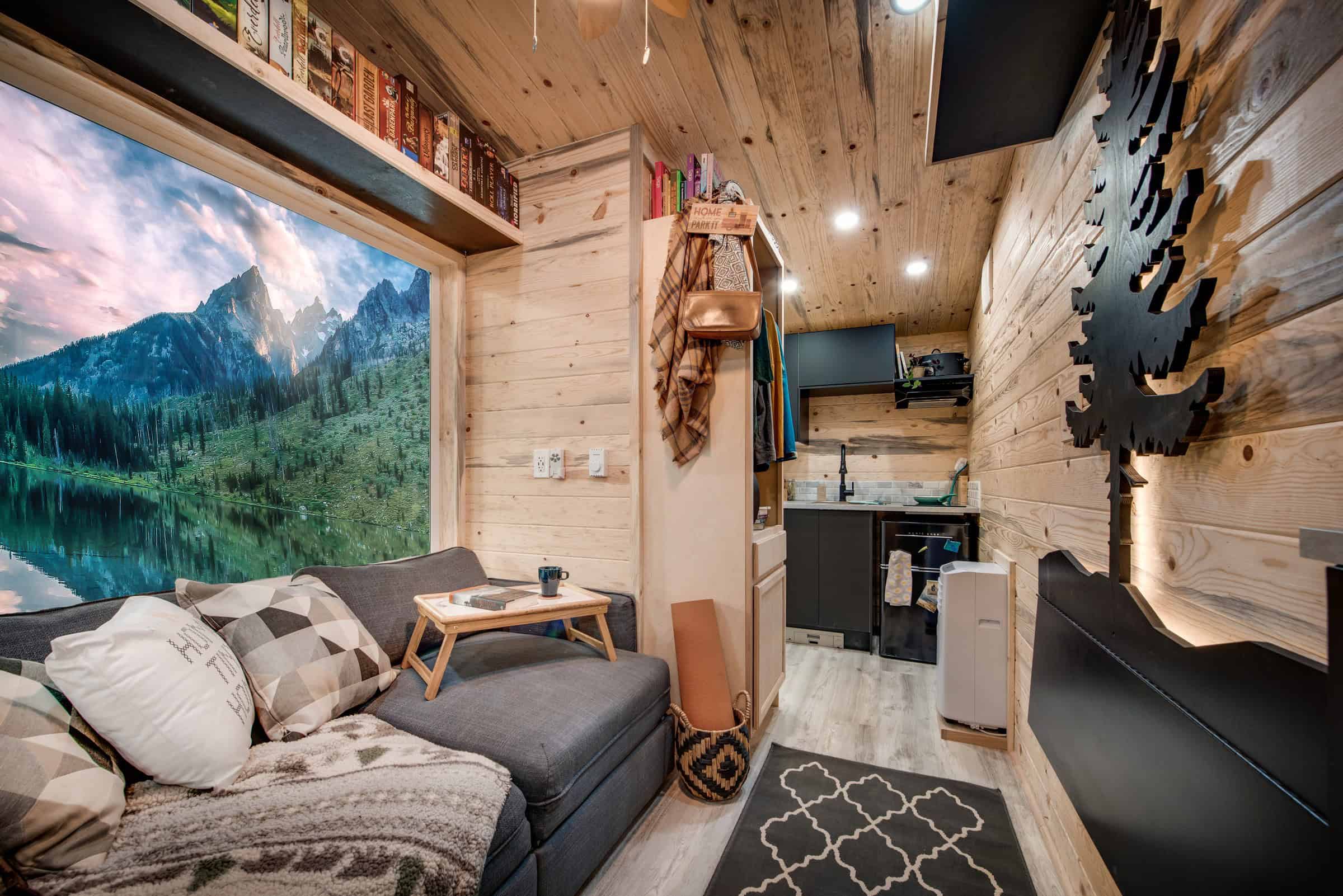 Traveler living toward kitchen: knotty pine, grey sleeper sofa, backlit tree, wardrobe cabinet, compact kitchen with dark cabinets at the far end