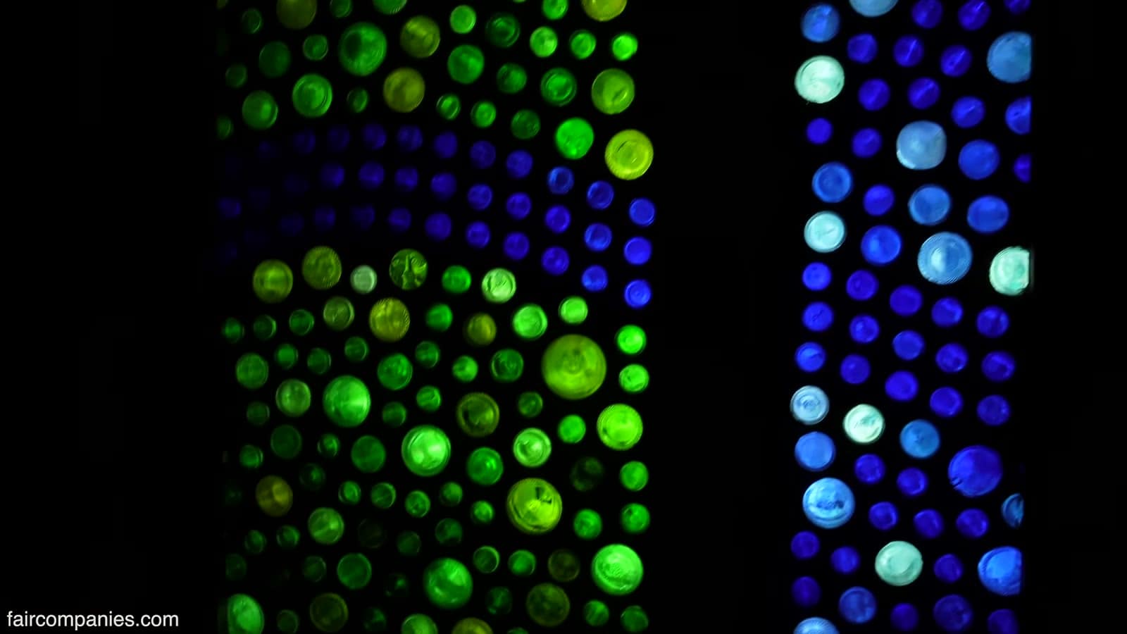 Backlit wall of recycled glass bottle bottoms glowing bright green and blue against darkness