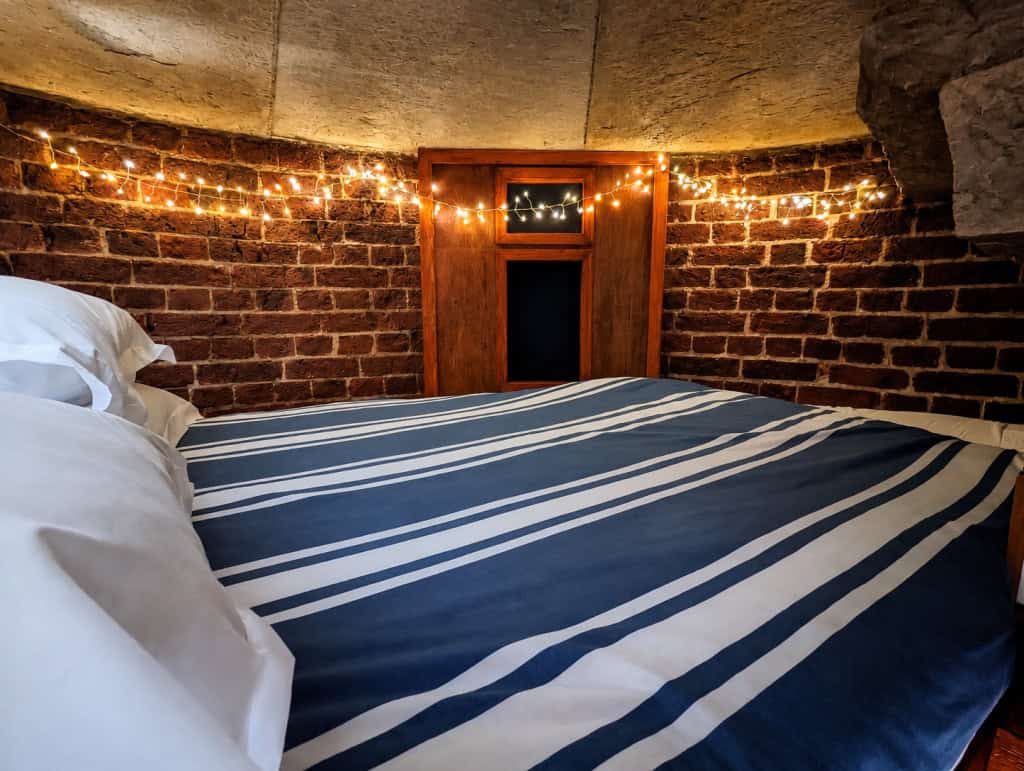 Keeper's Loft double bed with blue and white striped bedding against the curved brick wall of the lighthouse tower, with string lights