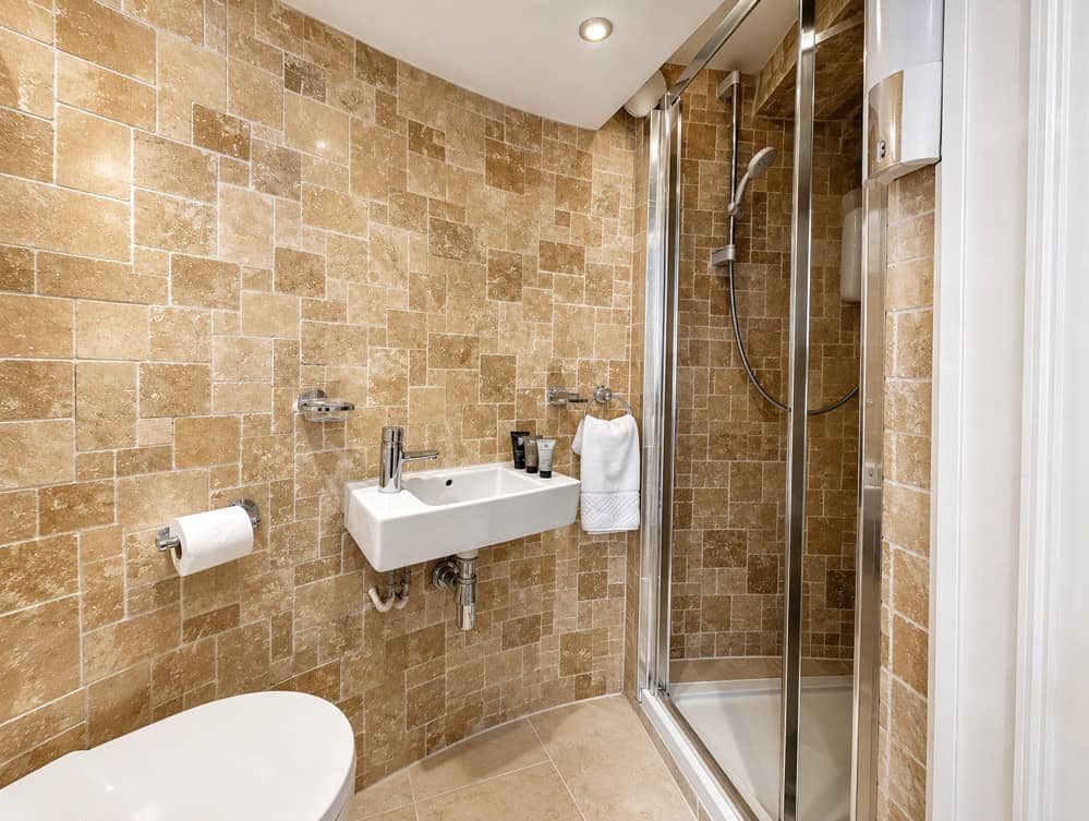 Keeper's Loft en-suite shower room with stone tile walls, small wall-mounted sink, toilet, and glass shower