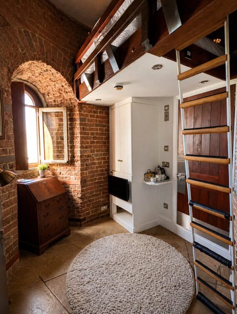 Keeper's Loft tower room: curved brick walls, arched window, antique desk, white built-ins, and a ladder up to the loft bed