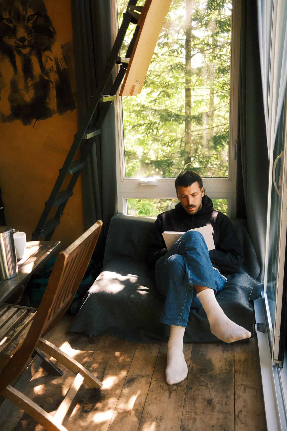 Living area with sofa at forest window and loft ladder inside Canopée tiny cabin