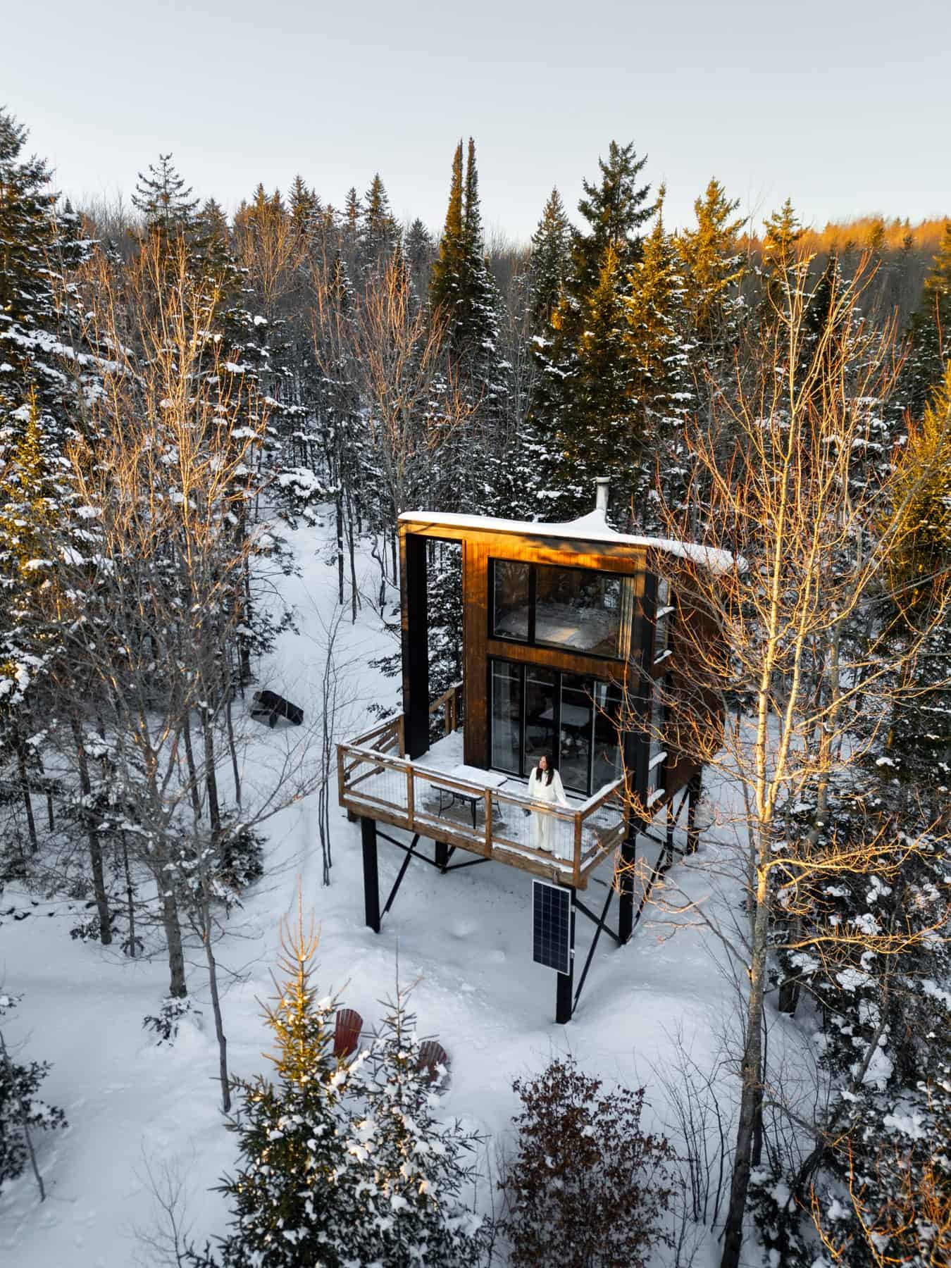 Winter exterior of Canopée stilt cabin in snow with deck figure and red chairs, Racine Eastern Townships