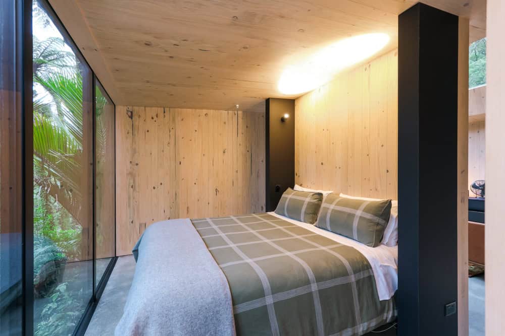 Bedroom with a plaid bed aimed at floor-to-ceiling forest glass