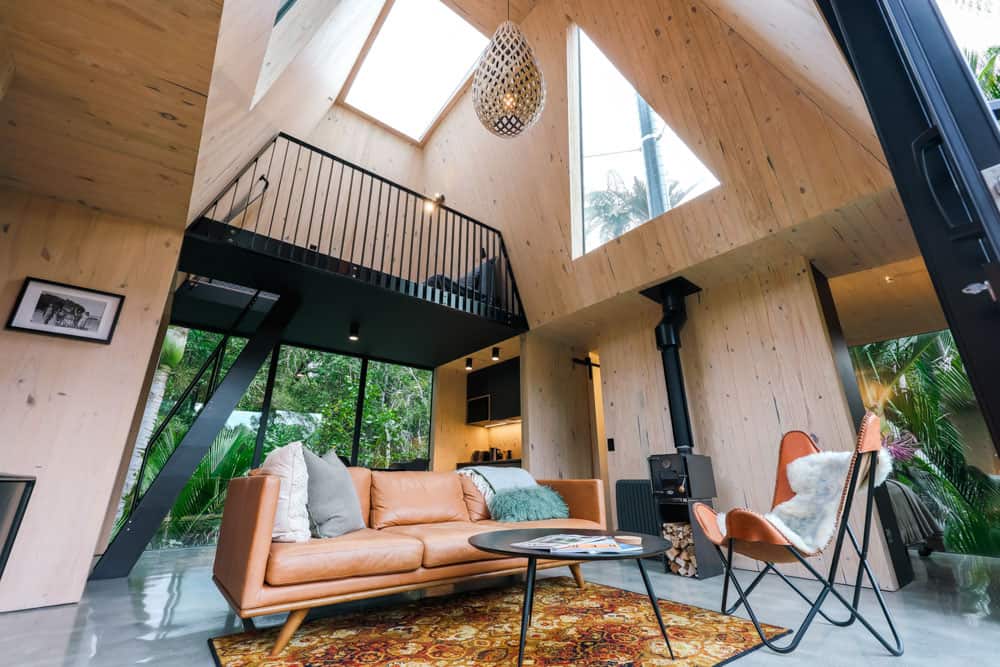 Vaulted light-wood living room with leather sofa, black stove, loft, and forest sliders