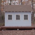 Pale blue 16x9 tiny cabin with a brown shingle roof, three white-trim windows, and a wood deck on concrete piers in a fall hardwood forest in rural Maine