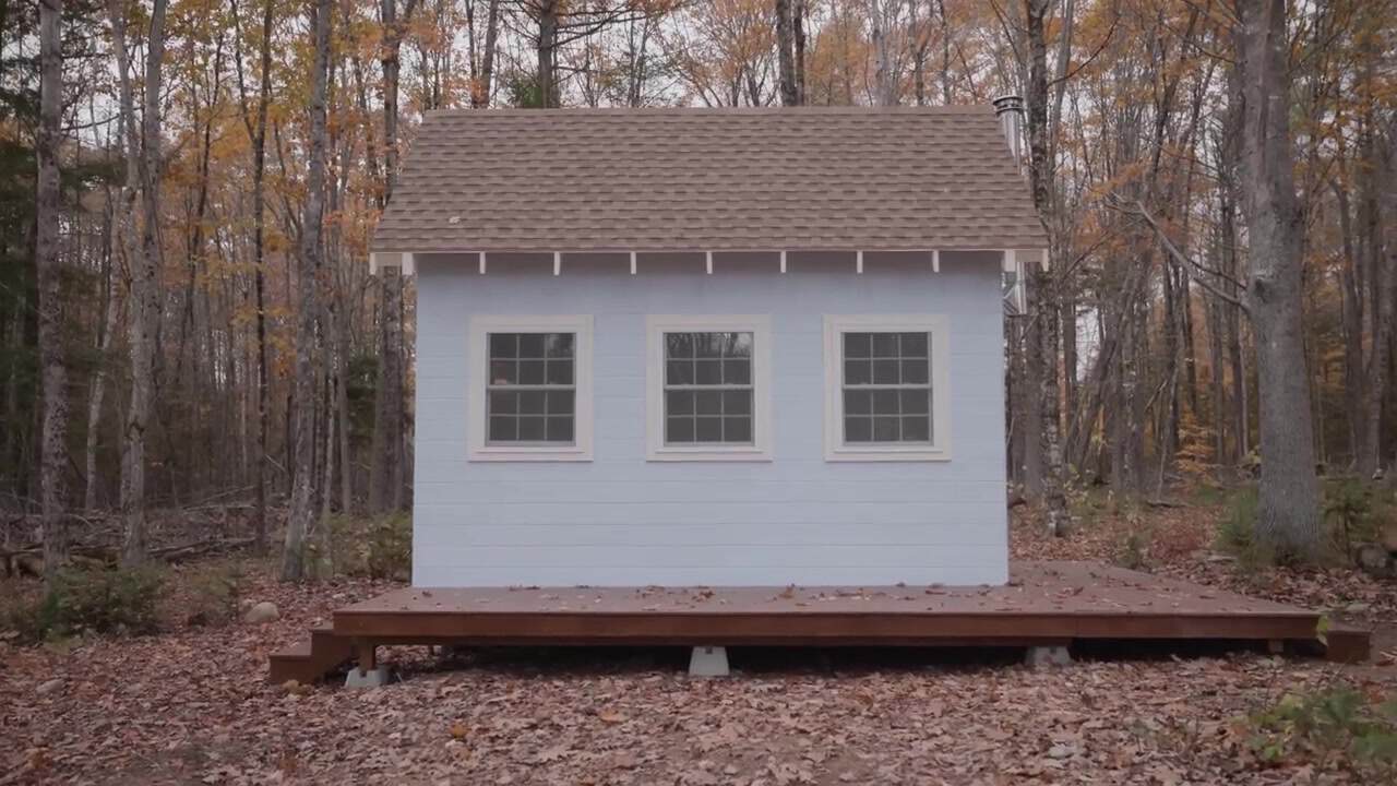 Pale blue 16x9 tiny cabin with a brown shingle roof, three white-trim windows, and a wood deck on concrete piers in a fall hardwood forest in rural Maine