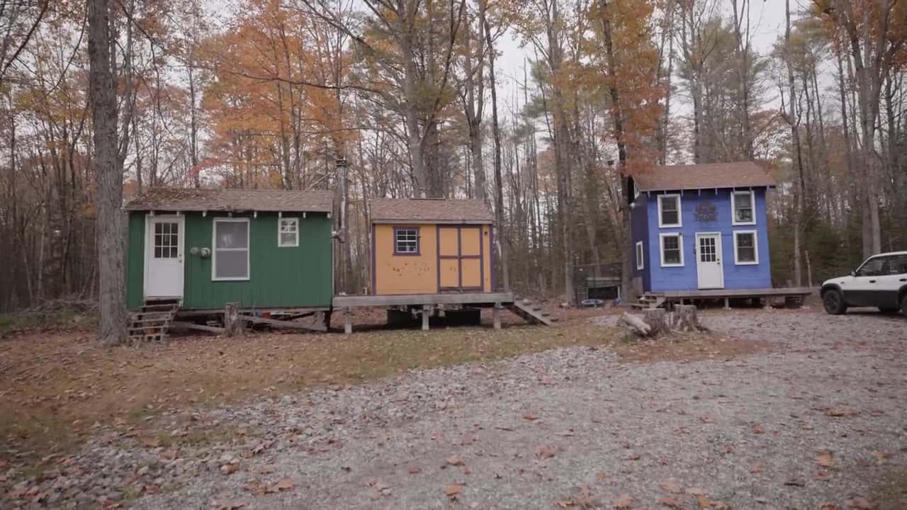 Three small off-grid cabins — green, yellow, and blue — on posts in a fall forest, part of Little Village on 40 acres in rural Maine
