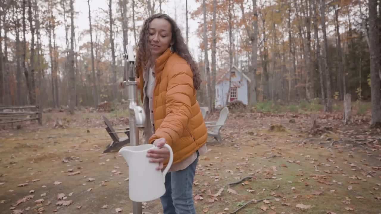 Bree at an outdoor hand-pump well filling a white pitcher, with her pale blue tiny cabin in the fall woods behind her