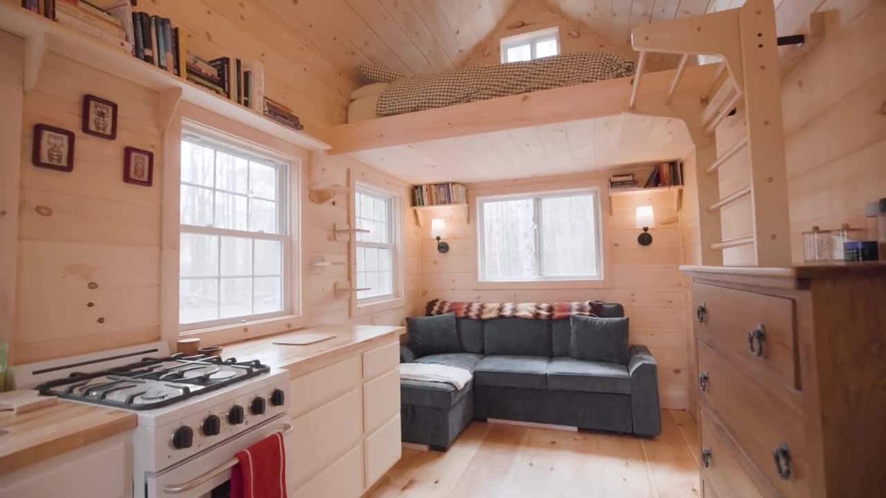 Looking from the kitchen toward a gray couch and a sleeping loft with gingham bedding, wall bars, and high pine ceilings