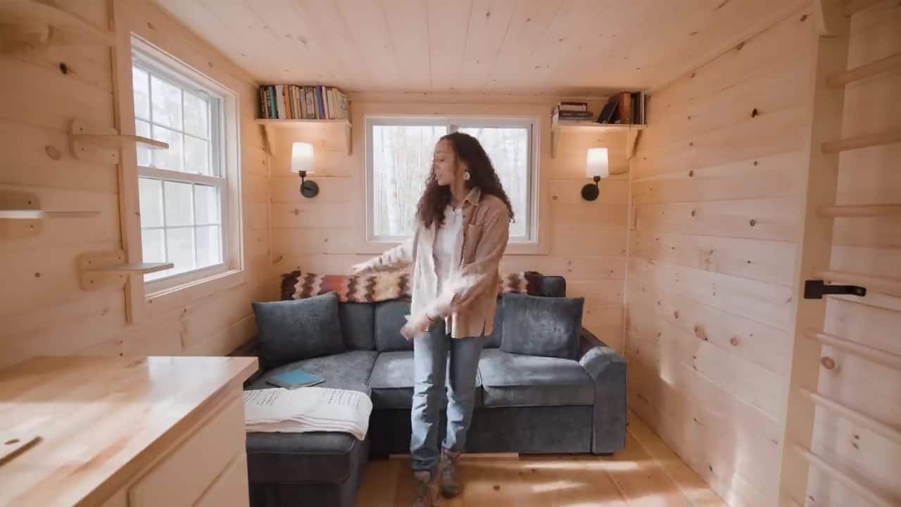 Bree in the living area of her tiny cabin, gray storage couch with a patterned throw, wall sconces, and windows to the woods