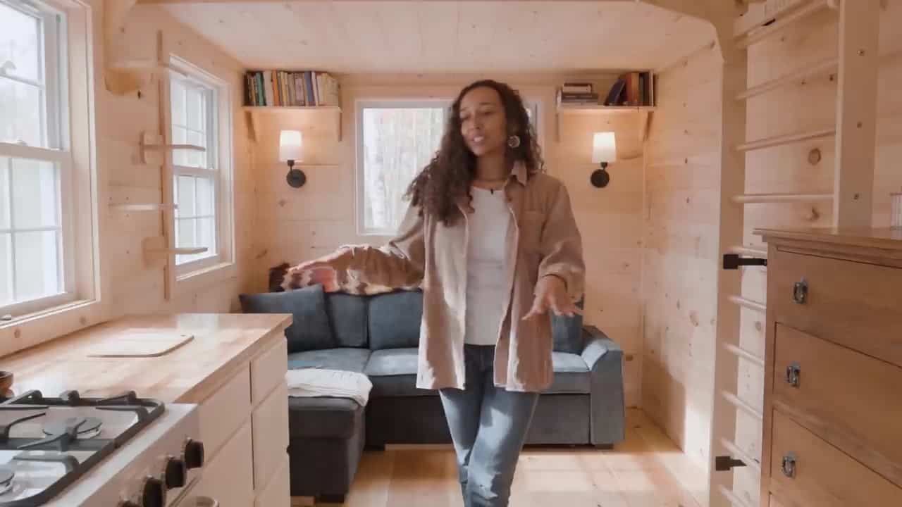 Bree in the living area of her 16x9 cabin with a gray couch, pine walls, a wood dresser, and wall bars used as a loft ladder and workout setup