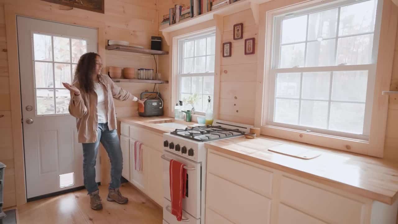 Compact off-grid kitchen with a white 20-inch propane stove, butcher block counters, preassembled white cabinets, open wood shelves, and a Jackery power station by the door