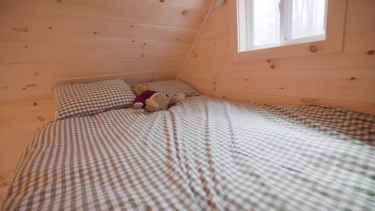 Full-size bed with gray gingham bedding and a stuffed bear under the pitched pine loft ceiling, with a small window to the woods