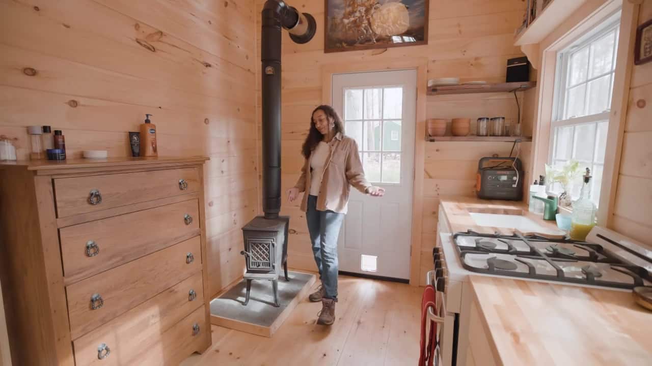 Bree standing beside a small black wood stove on a hearth pad, with a wood dresser to the left and the propane kitchen to the right