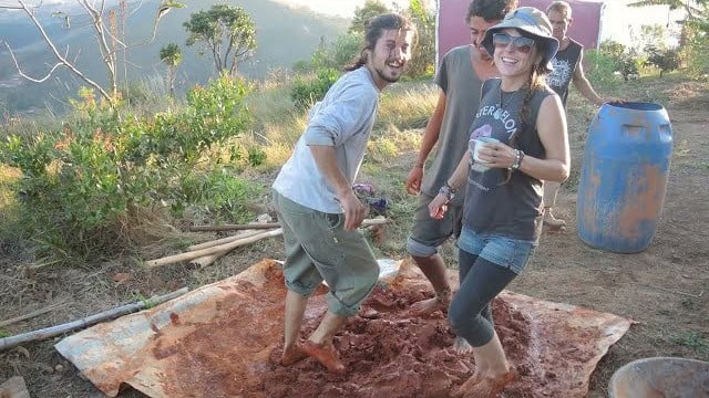 People mixing cob on a tarp on a grassy hillside, reddish earth pile and a blue barrel nearby