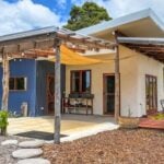 Cream earth-plaster house with a blue corrugated bay, timber-post porch, yellow shade sail, outdoor sink, and circular stepping stones near Denmark, Western Australia