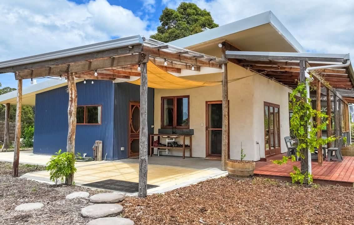 Cream earth-plaster house with a blue corrugated bay, timber-post porch, yellow shade sail, outdoor sink, and circular stepping stones near Denmark, Western Australia