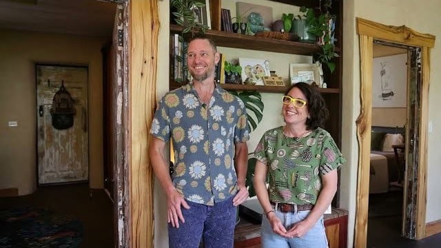 Brenna and Charlie standing between live-edge timber doorways in front of a built-in bookshelf, earth-plaster walls, a bed visible through the right door