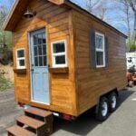 Cedar 7x14 cottage on a dual-axle trailer with a blue door, flower boxes, porch light, and wooden steps in Brookfield, Connecticut