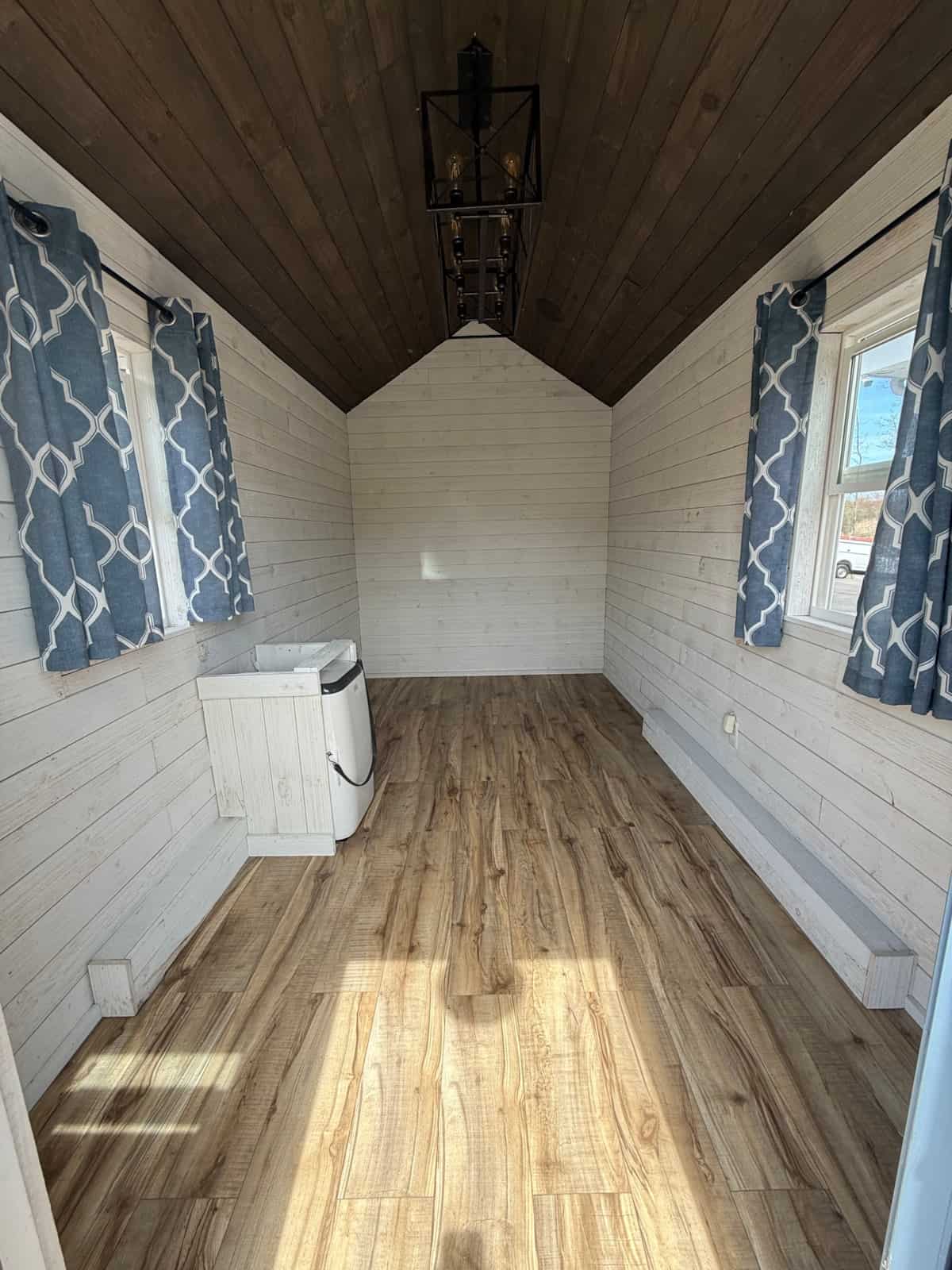 Open cottage shell with vaulted wood ceiling, blue geometric curtains, white shiplap, and a floor AC/heat unit