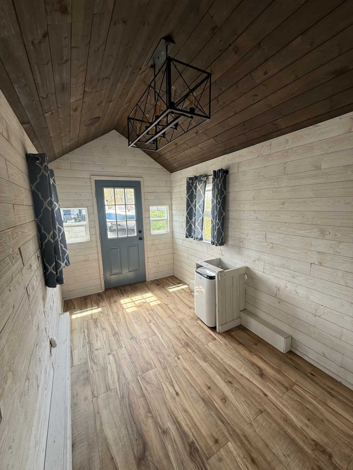 Open interior looking toward a blue door: white shiplap, wood-plank ceiling, vinyl plank floor, eight-bulb chandelier, AC/heat floor unit