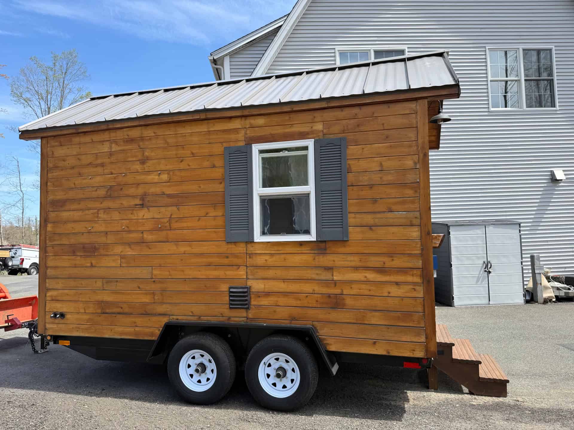 Side of the cedar cottage showing metal roof, shuttered window, and dual-axle trailer frame