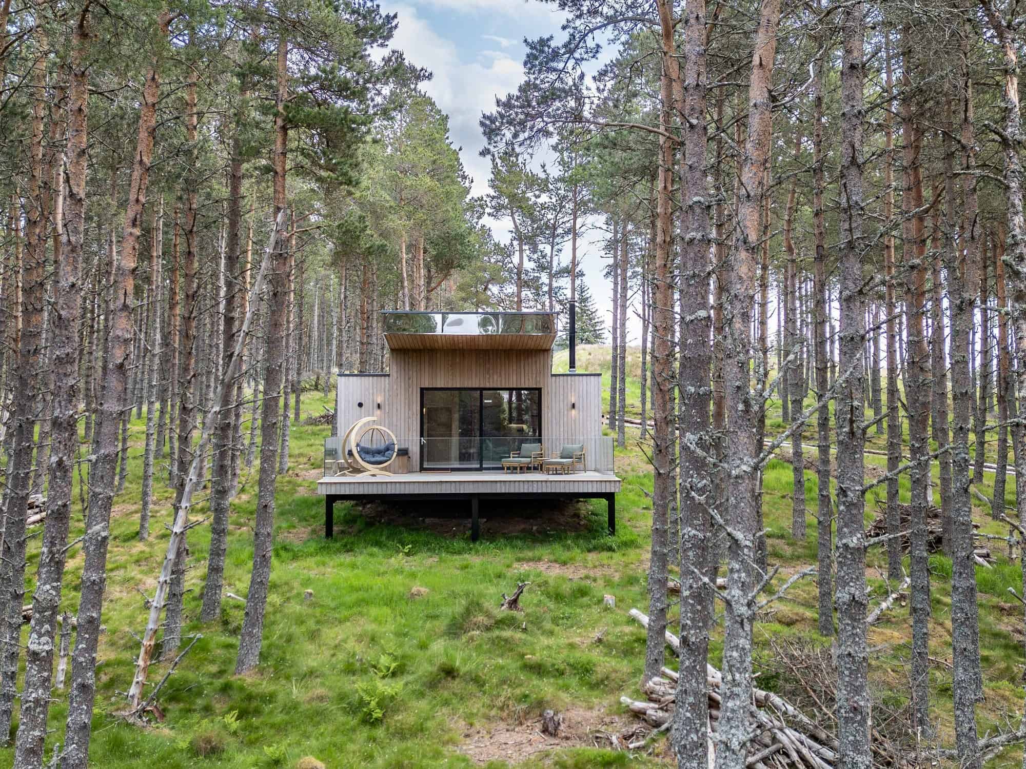 Wood cabin on stilts in a pine forest with floor-to-ceiling sliding glass, a glass-railed deck, a hanging lounger, and two lounge chairs
